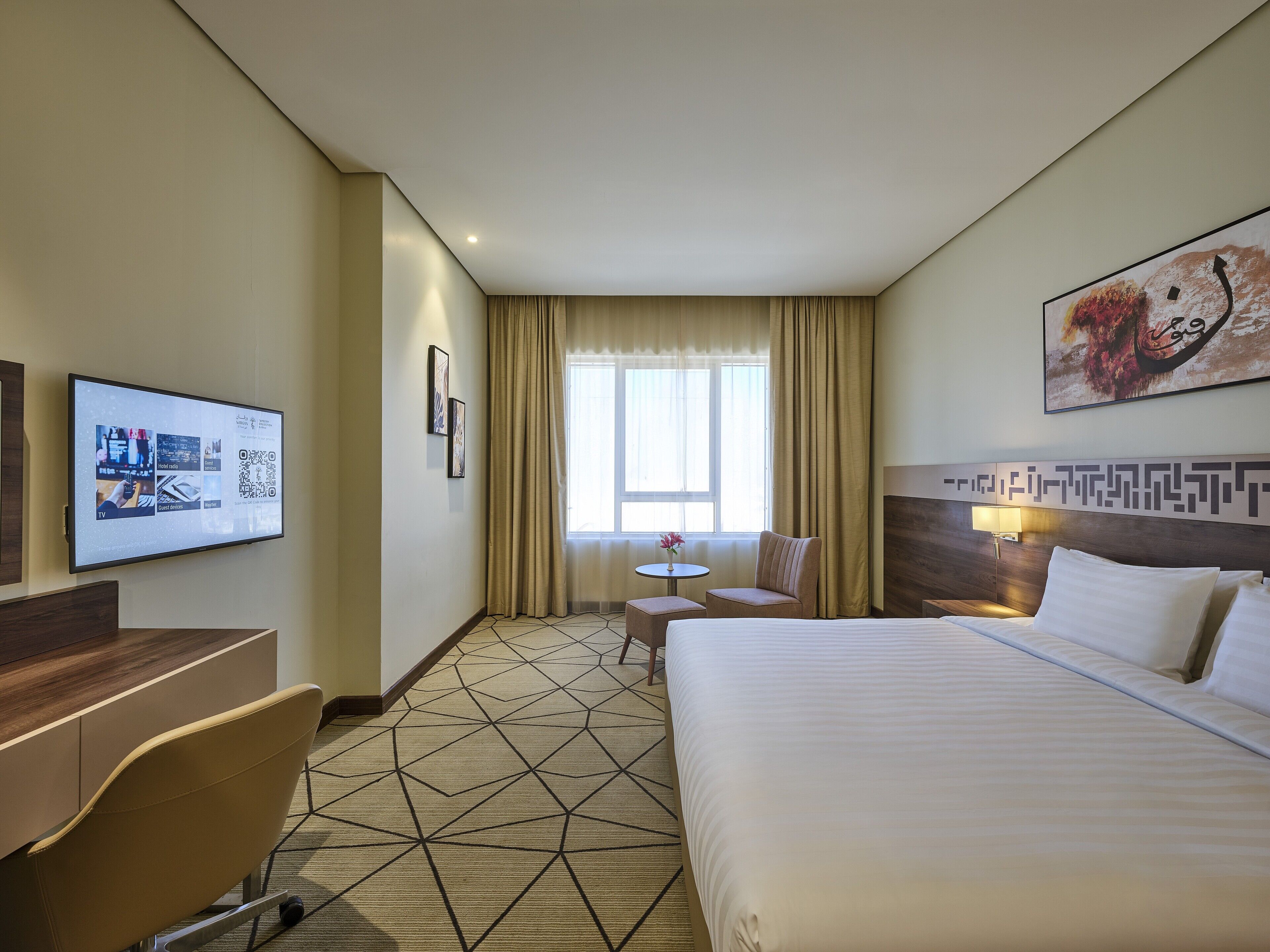 Quarto de hotel com cama, secretária, cadeira, mesa pequena e televisão fixada na parede.