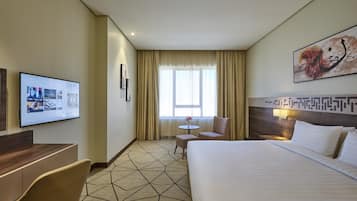 Quarto de hotel com cama, secretária, cadeira, mesa pequena e televisão fixada na parede.