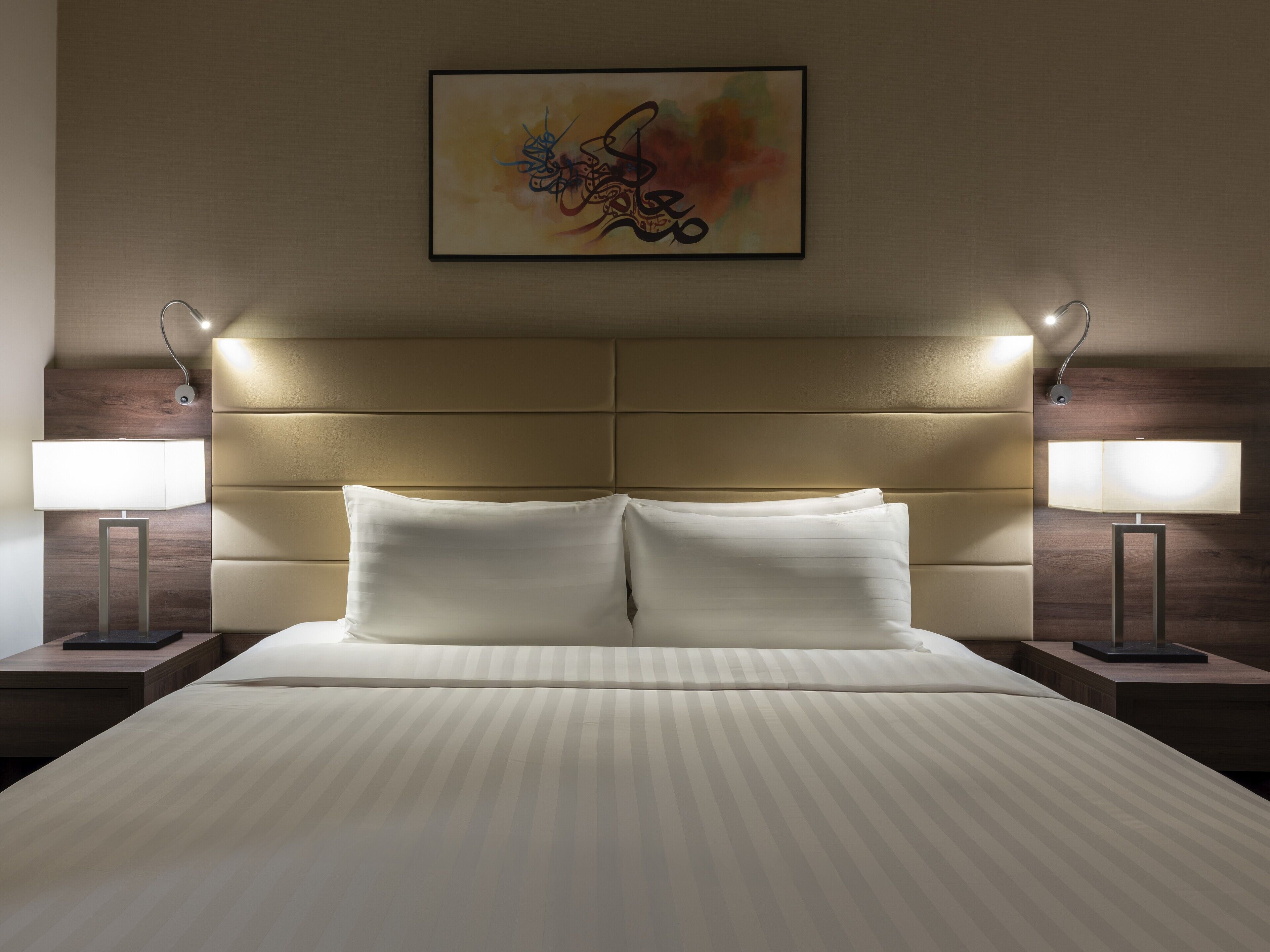 Una habitación de hotel con una cama grande, dos lámparas de noche, un cabecero y una obra de arte enmarcada en la pared.