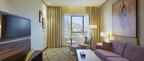 A hotel room with a flat-screen TV, a sofa, a coffee table, a chair, and a view of a mountainous landscape through a window.