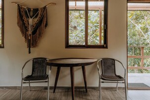 Una habitación con una mesa redonda y dos sillas de mimbre, un colgador de macramé en la pared y una ventana con vista a árboles.