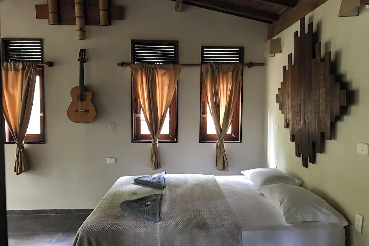 A room with a bed, a guitar, and three windows with curtains.