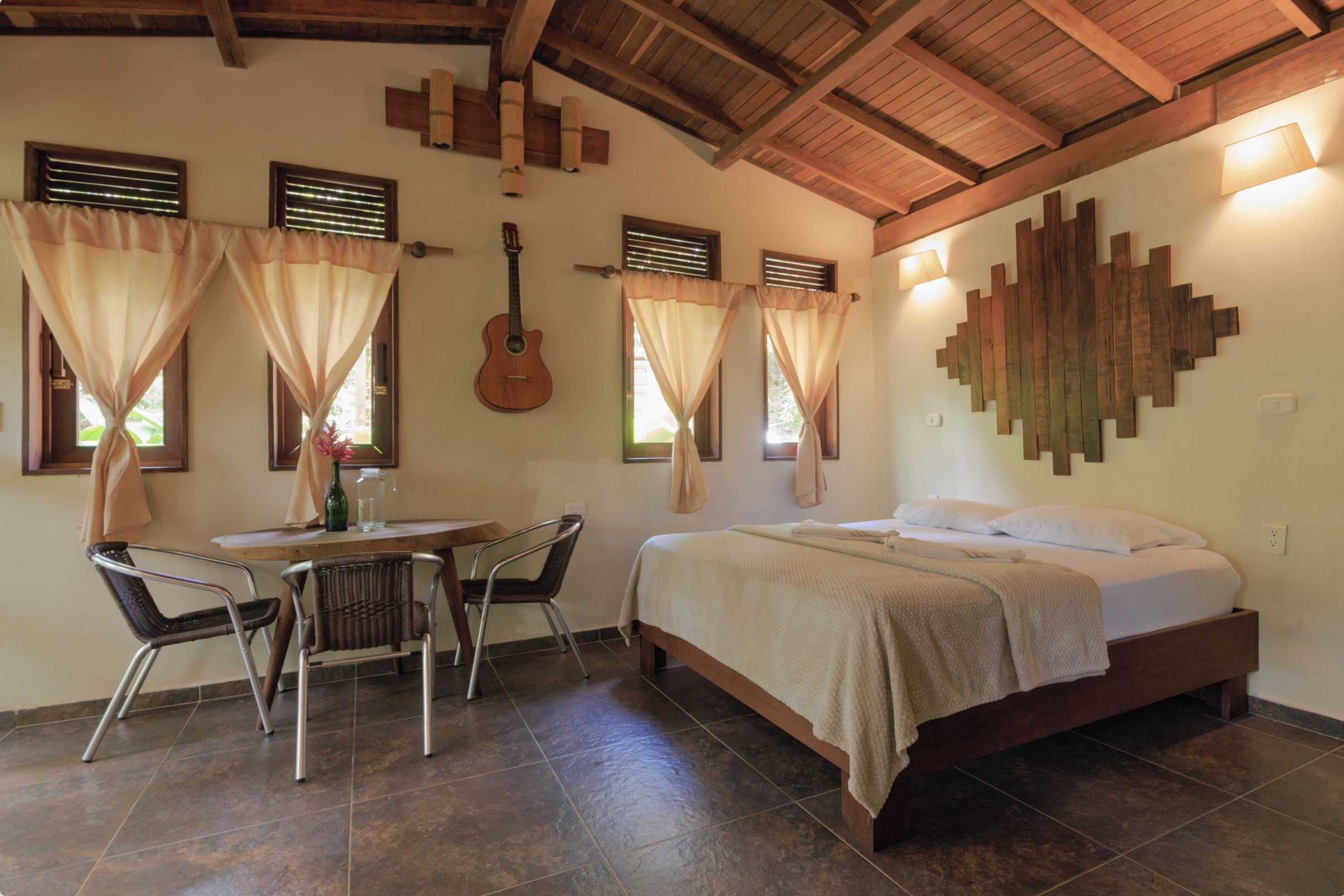 A bedroom with a bed, a guitar, and a dining area with chairs and a table.