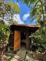 Una cabaña de madera con una sola puerta y un pequeño porche, rodeada de densa vegetación tropical.