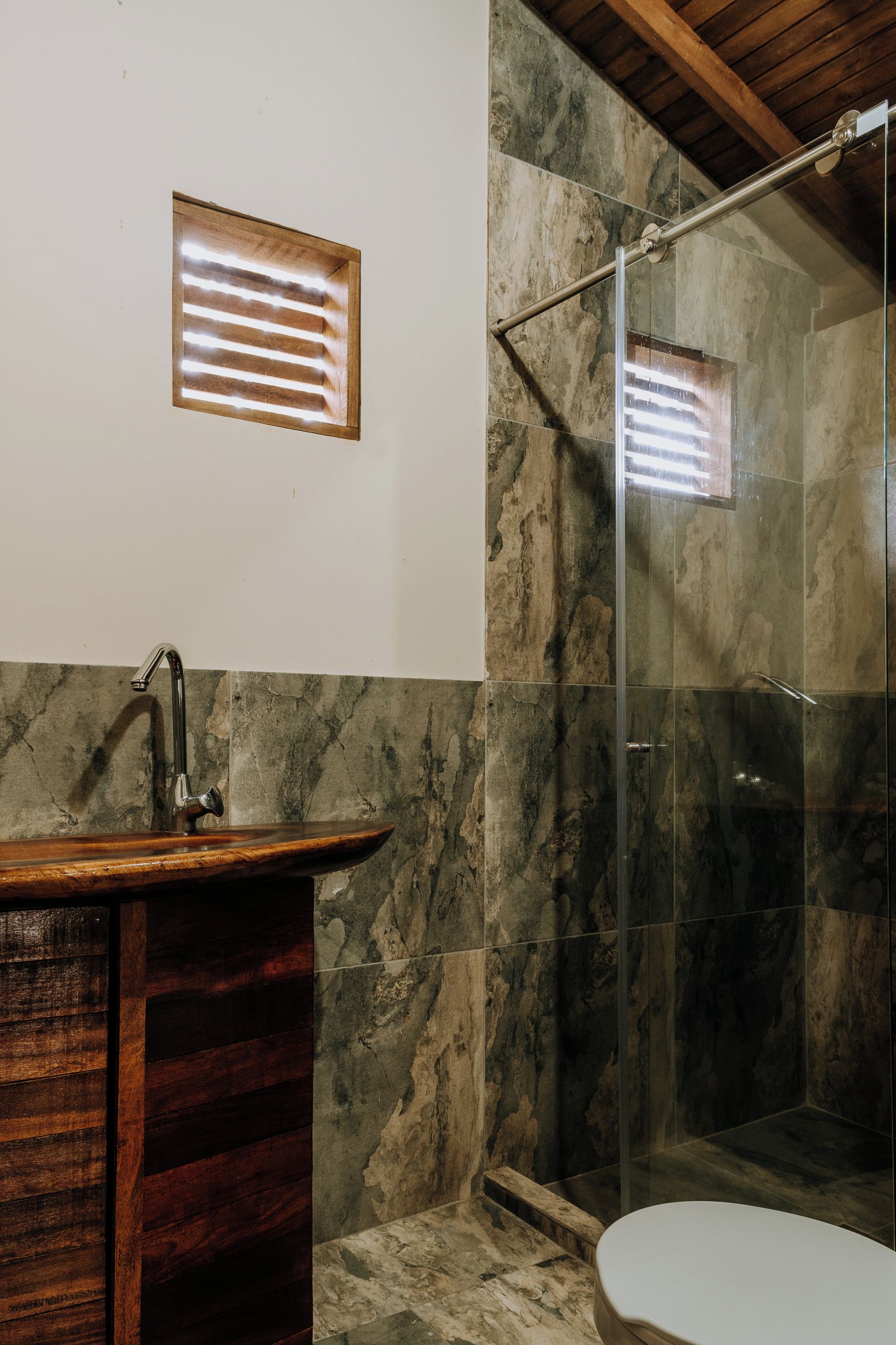 A bathroom with a wooden vanity, a glass shower enclosure, and a marble wall.