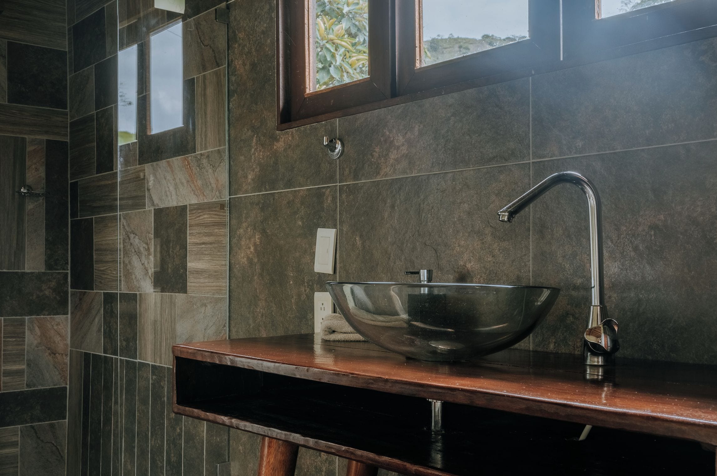 A modern bathroom with a large vessel sink, a wooden vanity, and a window with a view of greenery.