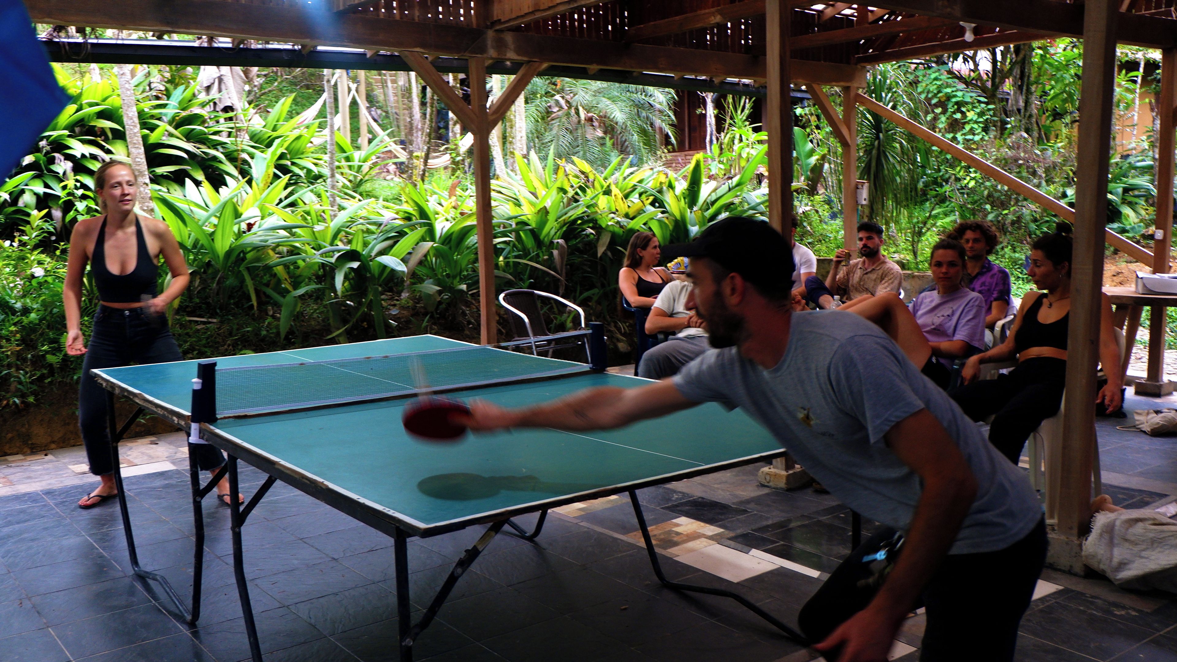 An outdoor table tennis game with a crowd watching, surrounded by lush greenery.