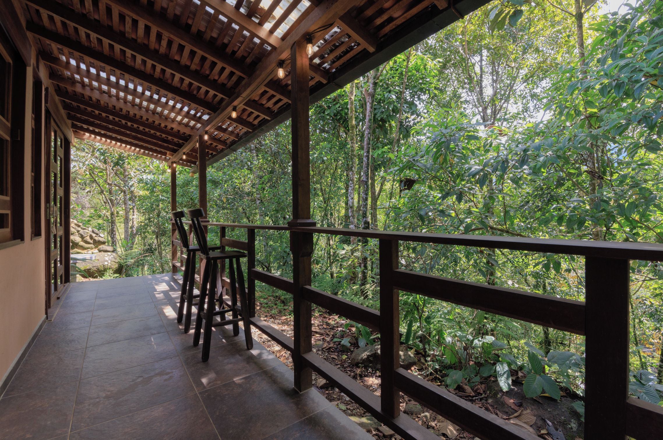 Un espace extérieur abrité, doté d’une pergola en bois, d’un sol carrelé et offrant une vue sur une forêt dense.