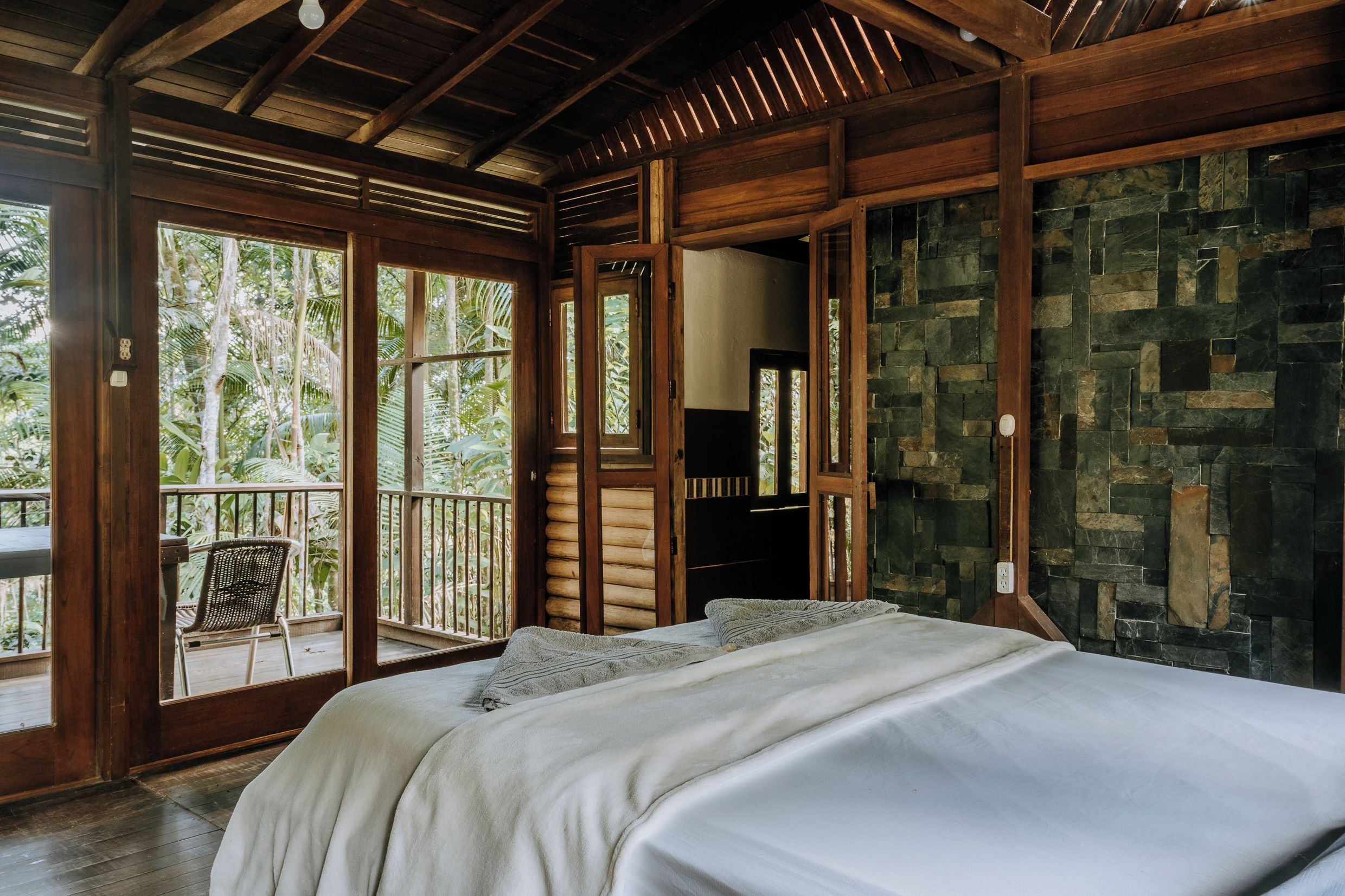 Una habitación con cama, techo de madera, pared de piedra y balcón con una silla.