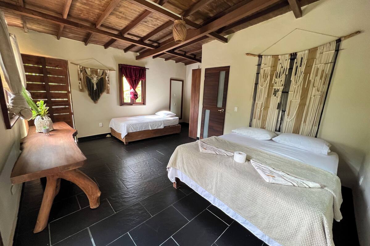 A room with a wooden ceiling, two beds, a small table with a plant, and a window with curtains.