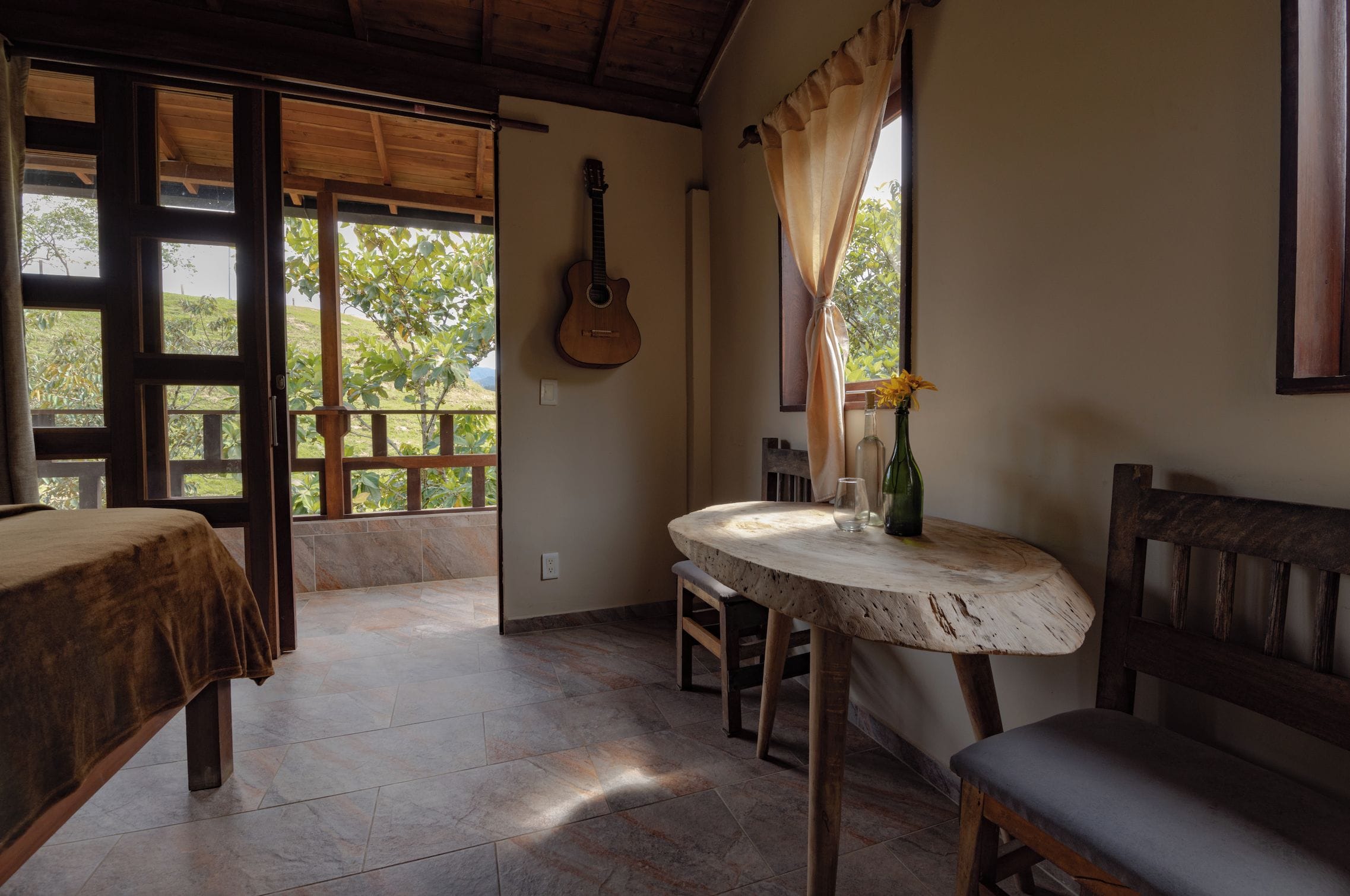 A room with a wooden guitar, a round table with a bottle and glass, and a bench.