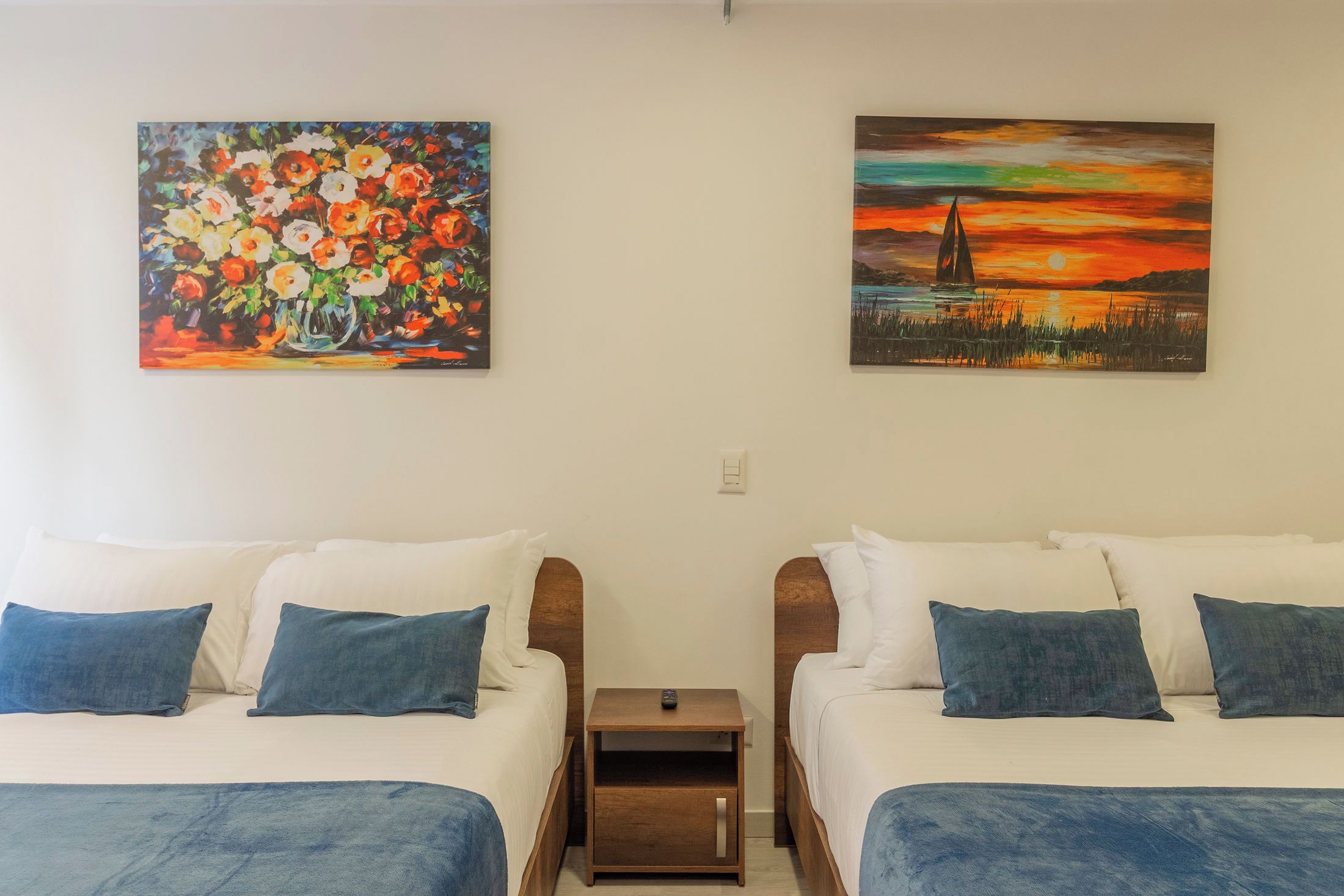 A hotel room with two beds, each with a blue pillow, a small wooden nightstand, and two paintings on the walls.