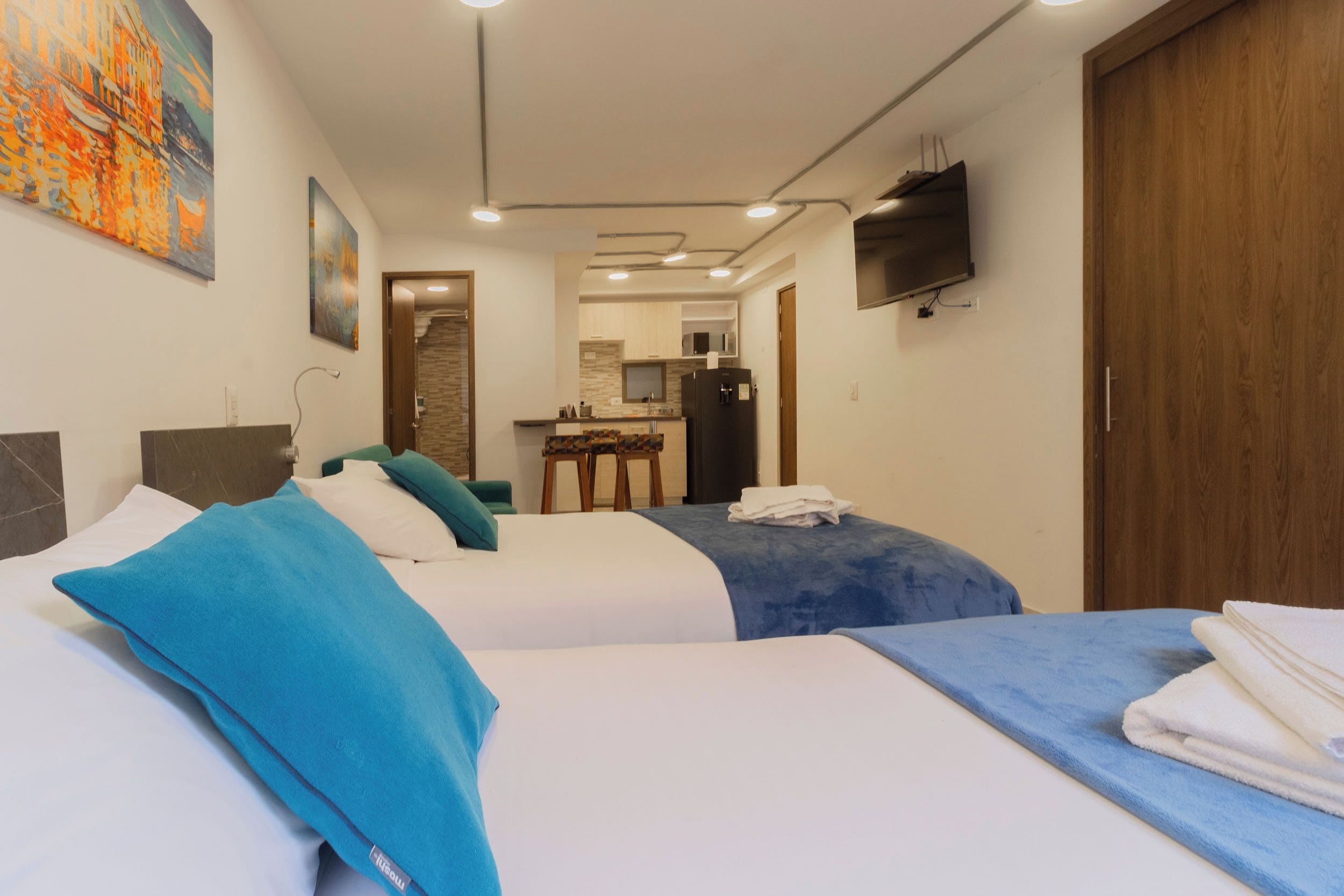A hotel room with two beds, a kitchenette, a TV, and artwork on the walls.