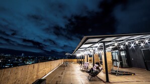 A rooftop terrace with a view of the city at night, featuring a covered seating area with chairs and a table, a treadmill, and a brick wall.