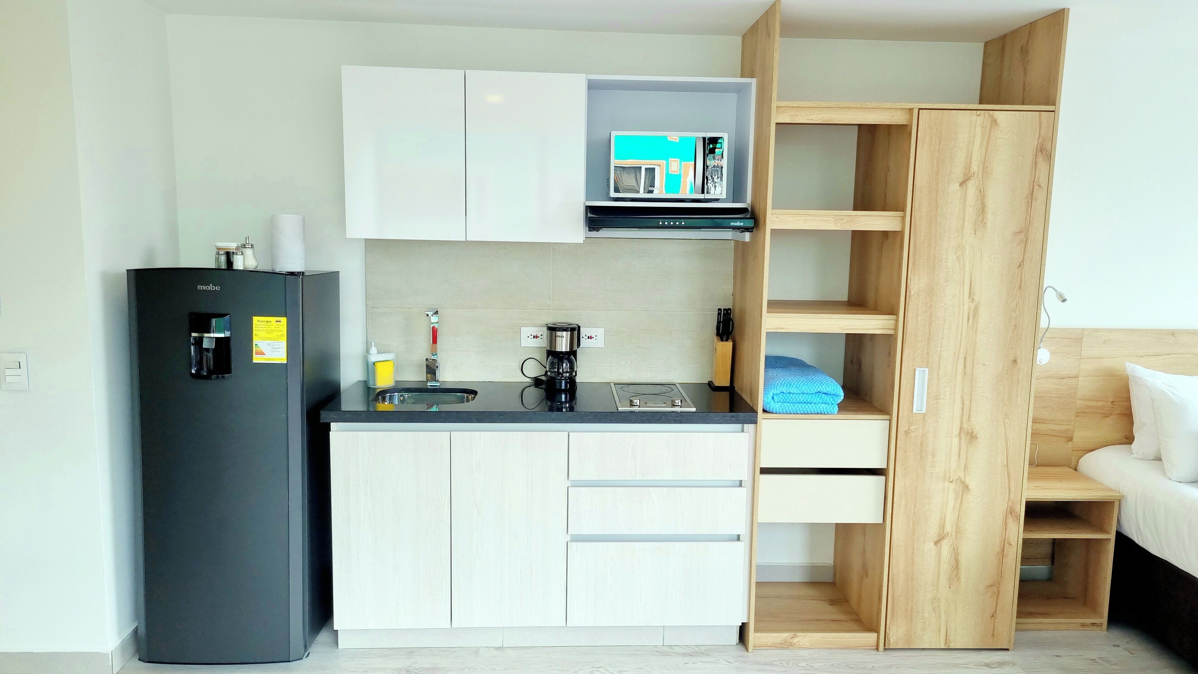 A compact kitchenette with a black refrigerator, a coffee maker, and a microwave. There is a wooden wardrobe with a bed in the adjacent room.