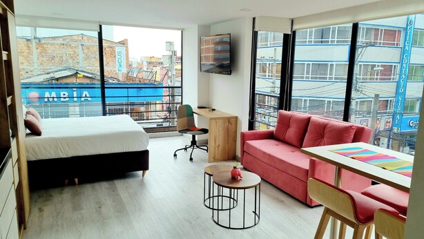 A modern hotel room with a bed, a red sofa, a small table, and a TV mounted on the wall.