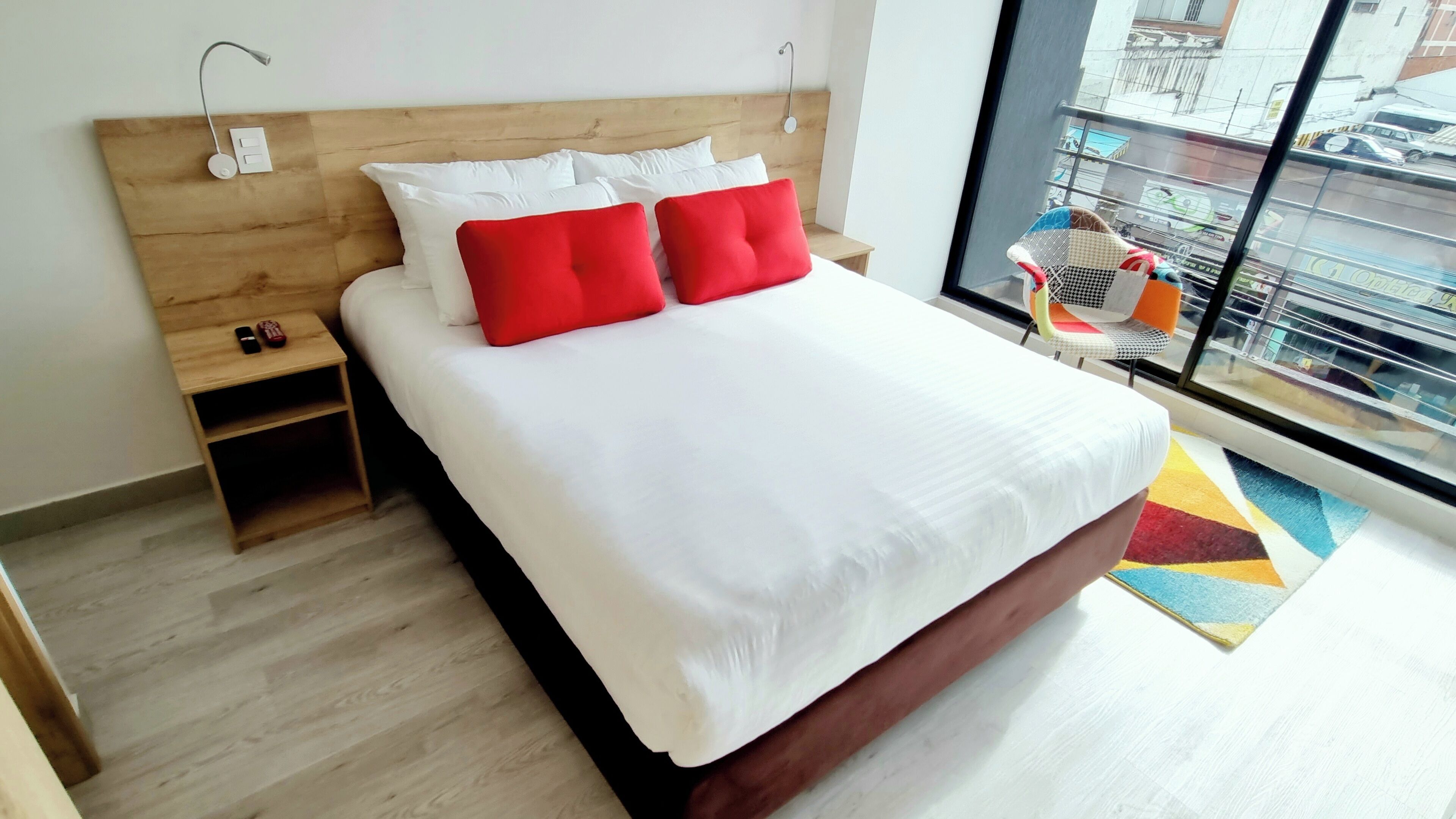 A hotel room with a large bed, wooden headboard, white bedding, and a view of the street through a large window.