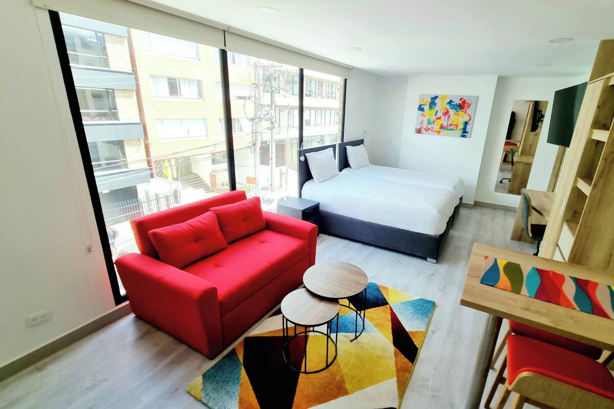 Une chambre d’hôtel moderne avec un grand lit, un canapé rouge, une petite table ronde et un tapis coloré.