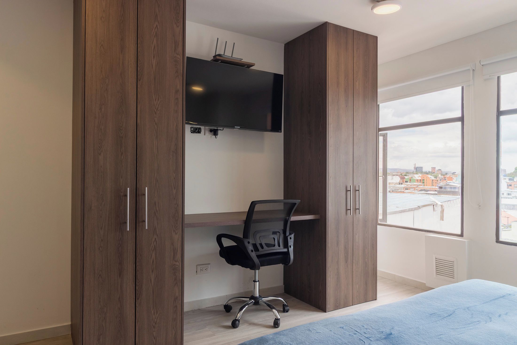 A bedroom with a bed, a desk with a chair, a TV mounted on the wall, and a window with a city view.