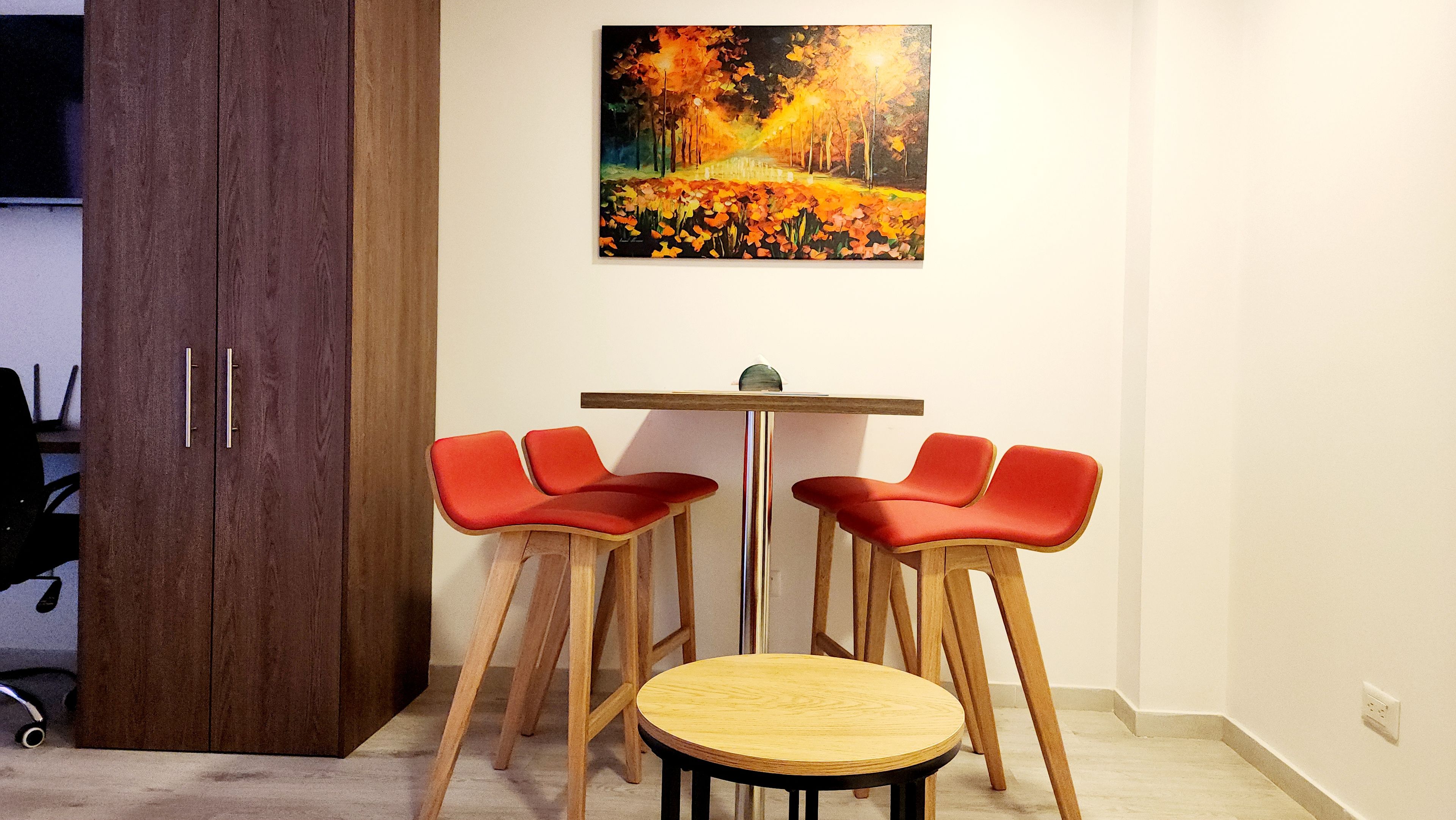 Een kamer met een houten tafel, vier rode stoelen, een schilderij aan de muur en een gesloten houten garderobe.