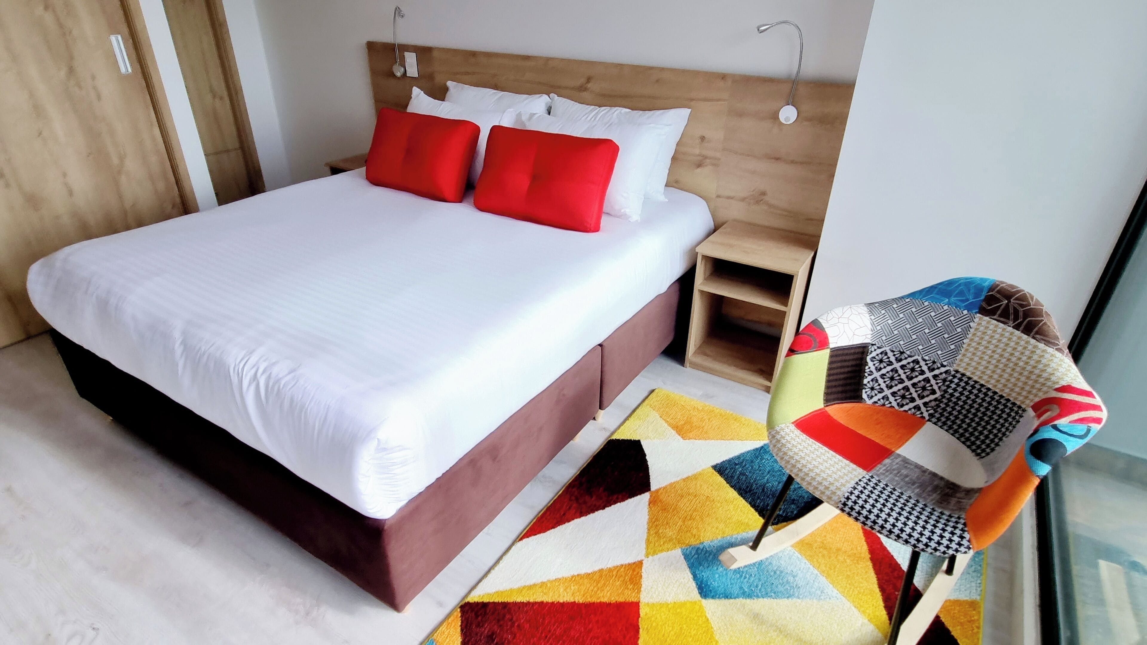 A modern hotel room with a large bed, a wooden headboard, a bedside table, and a colorful chair.
