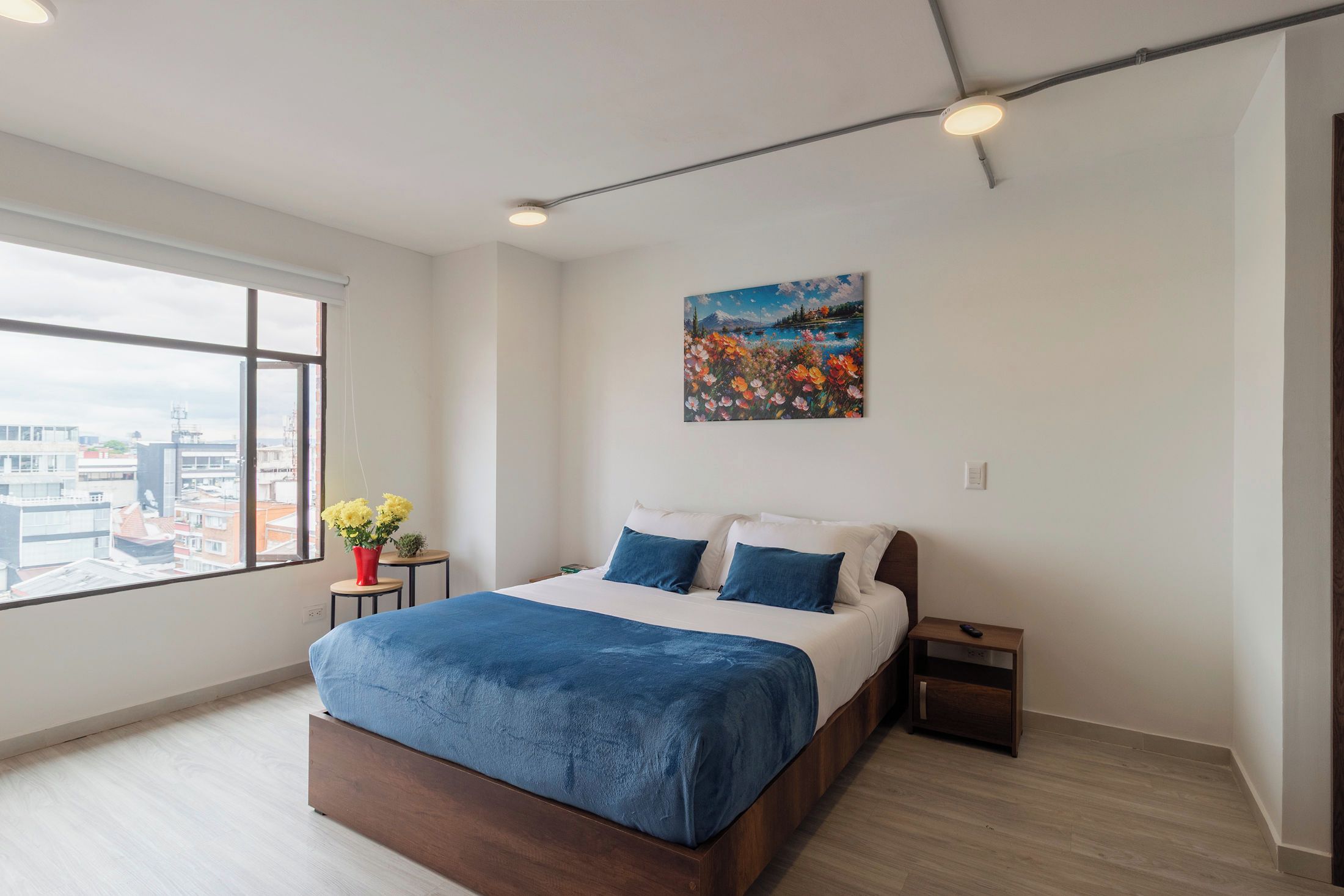 A modern bedroom with a large bed, a painting, a window with a view, and a small table with flowers.