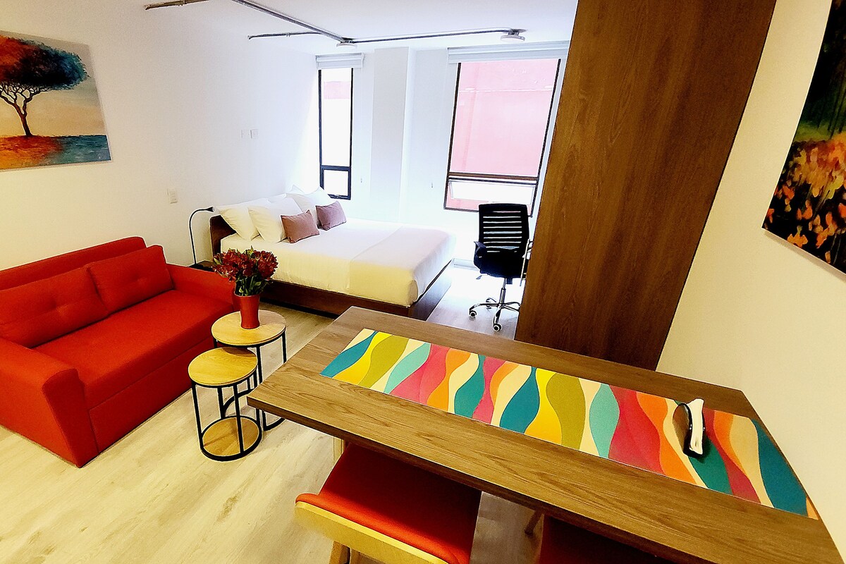 Een moderne kamer met een rode bank, een houten eettafel, een klein rond tafeltje met een vaas, een bed met wit beddengoed en een raam met zonwering.