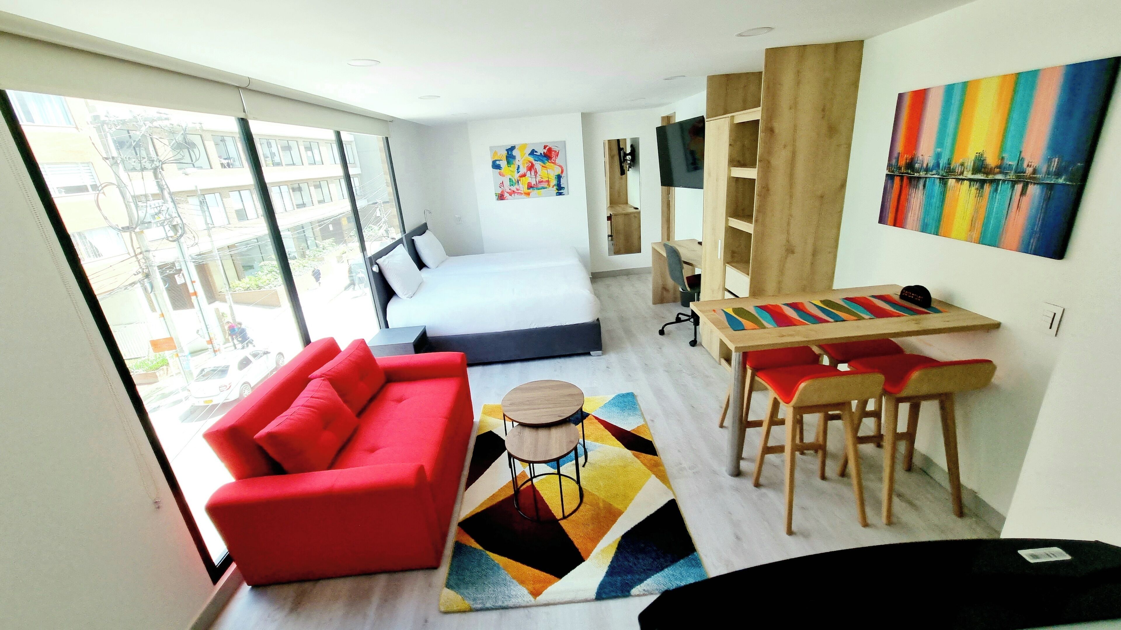 Una habitación de hotel moderna con una cama grande, un sofá rojo, un escritorio de madera con sillas y un cuadro abstracto colorido.