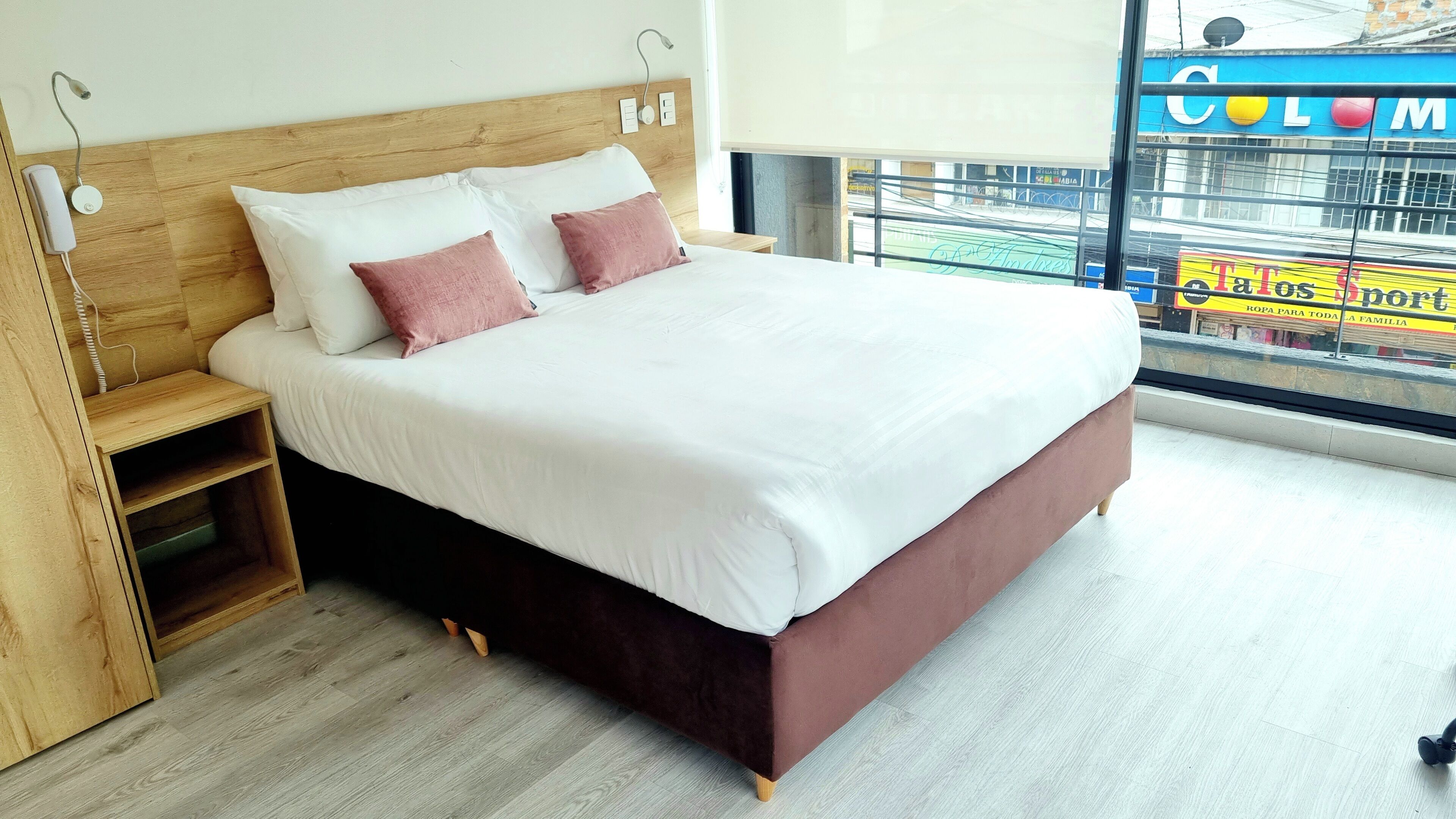A modern hotel room with a wooden headboard, a bed with white and pink bedding, a small wooden nightstand, and a view of a colorful street through the window.
