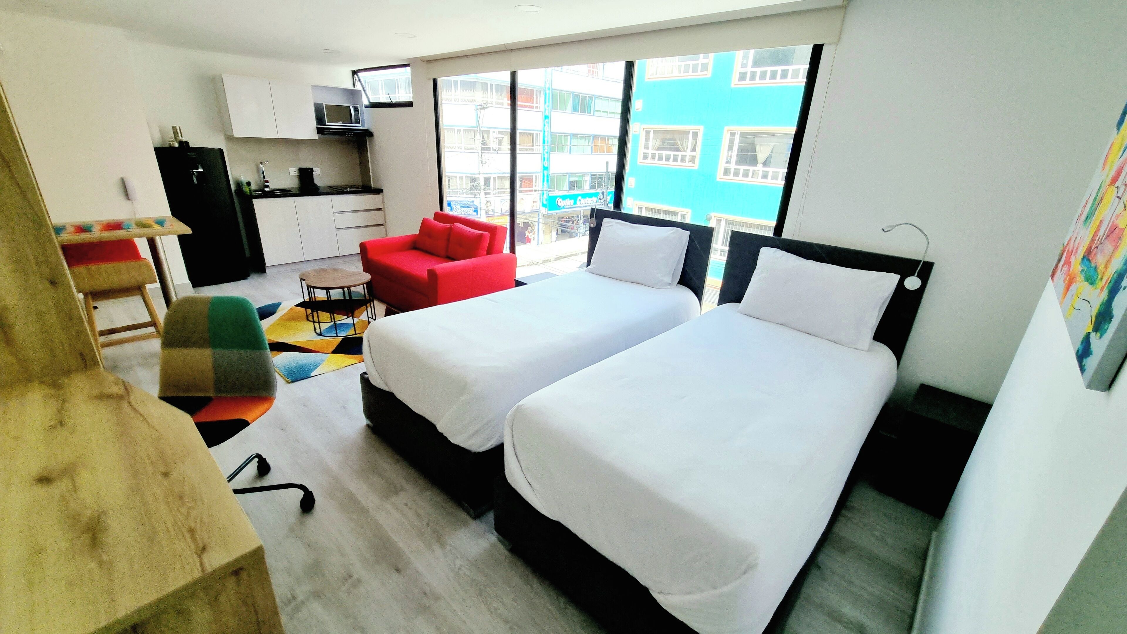 A small hotel room with two beds, a kitchenette, a red chair, and a view of a building through the window.