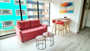 A modern living room with a red sofa, two round tables, and a small desk with red chairs. A colorful painting is on the wall, and the room has large windows with a view of a building.