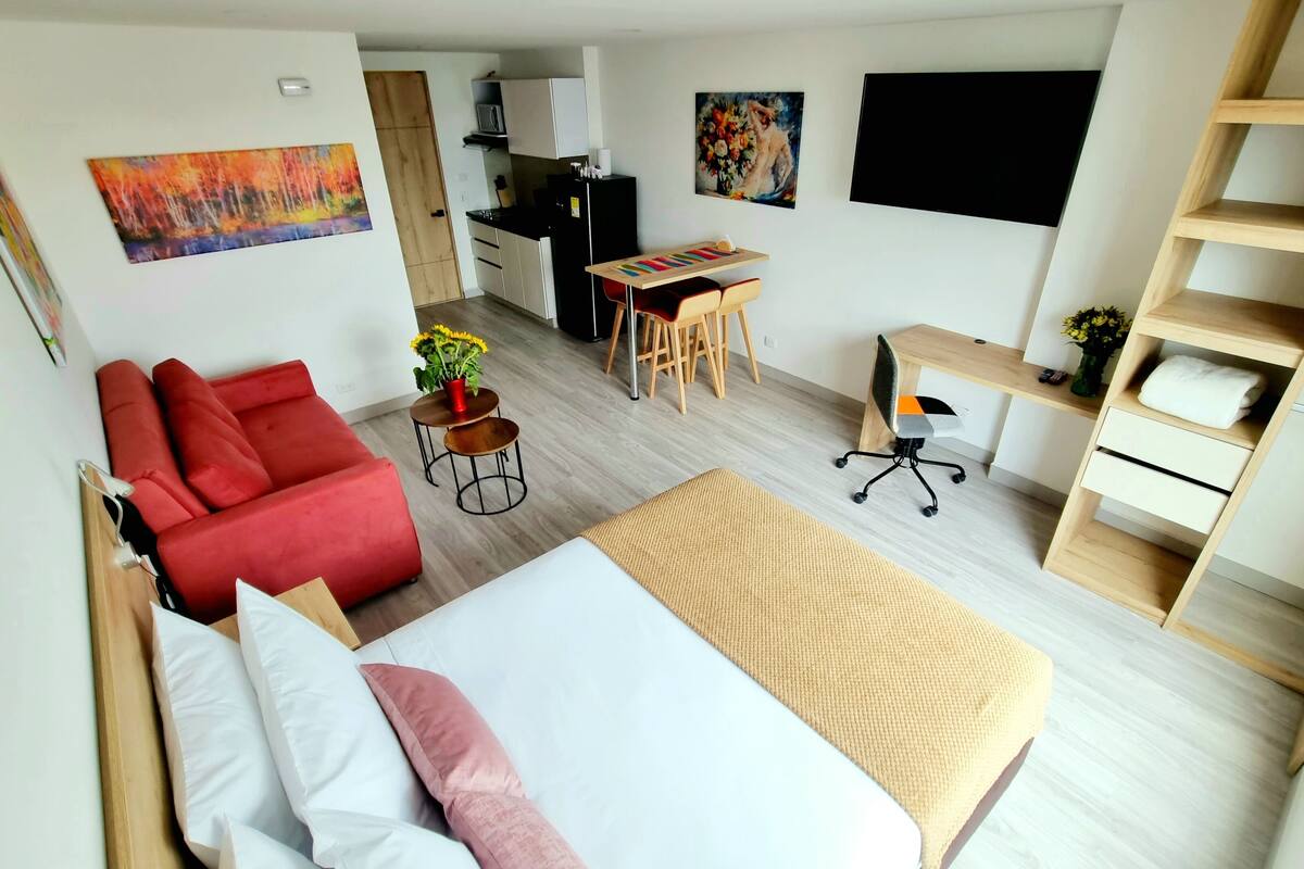 Une chambre moderne et bien éclairée, dotée d’un grand lit, d’un canapé rouge, d’un bureau avec une chaise et d’un téléviseur fixé au mur.