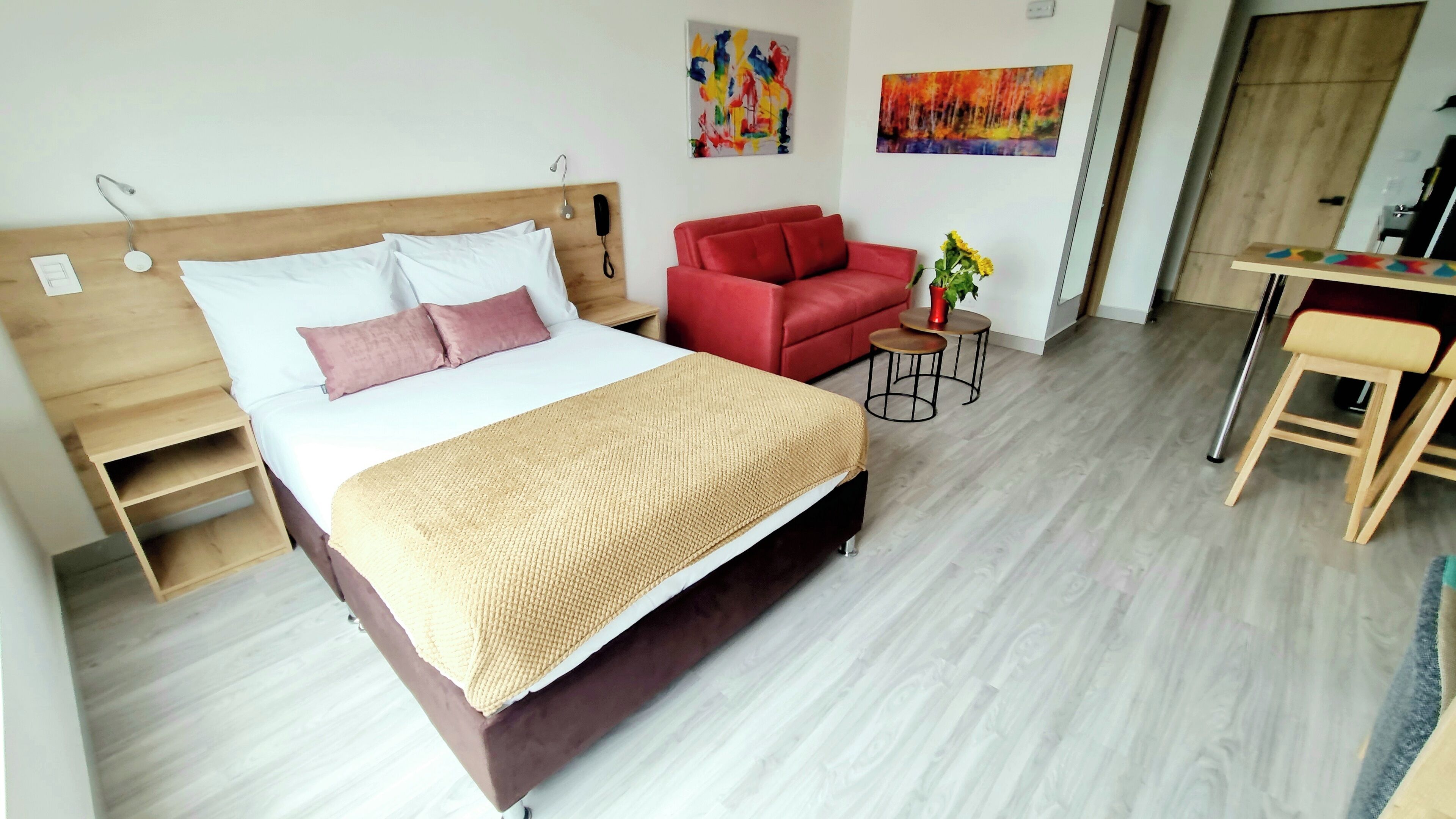 A hotel room with a bed, a red sofa, a small table with a vase, and a desk with a chair.
