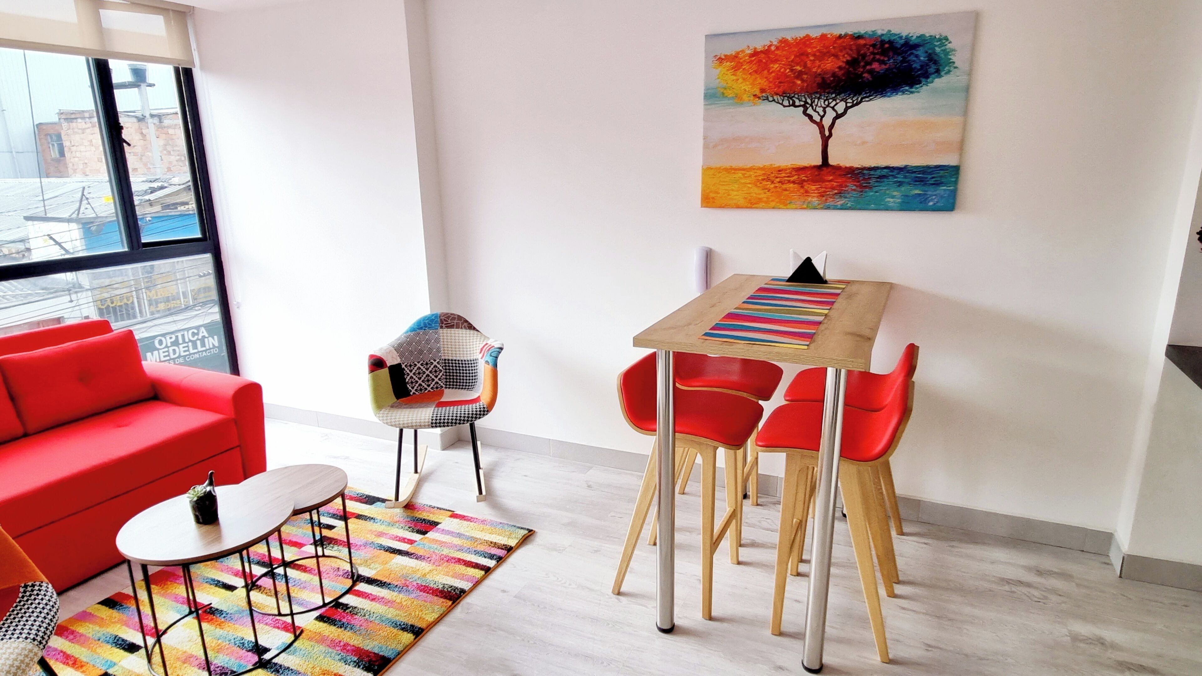 A modern living room with a red sofa, a small round table, and a colorful rug. A painting of a tree is on the wall, and a window offers a view of buildings outside.