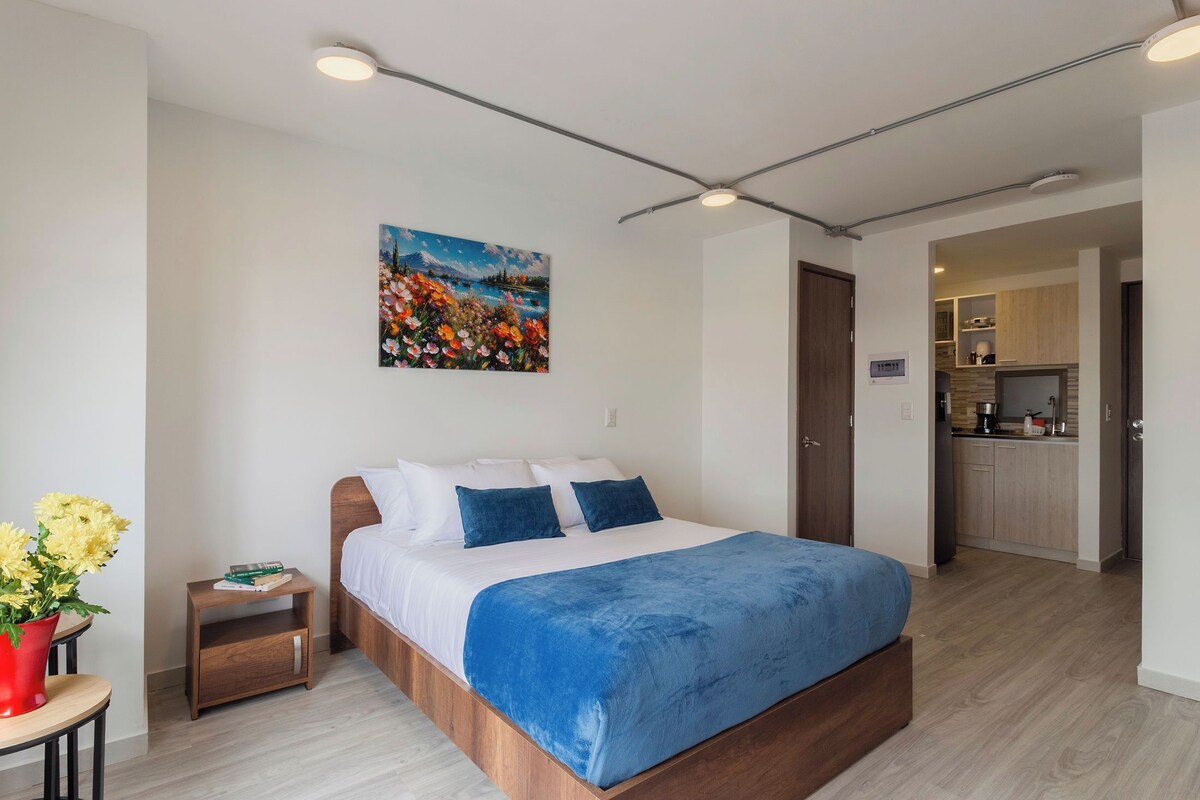 A modern bedroom with a wooden bed, a painting of a landscape, a small bedside table with books, and a vase of flowers.