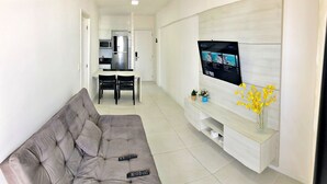 A living room with a grey sofa, a TV mounted on a white wall, a dining area with a table and chairs, and a kitchen with appliances. - Vacanze - Time (Maceió)