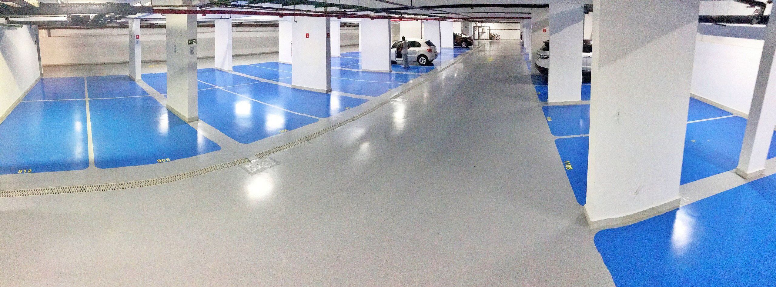 Un estacionamiento con pisos azules y blancos, pilares blancos y una persona cerca de un automóvil blanco.