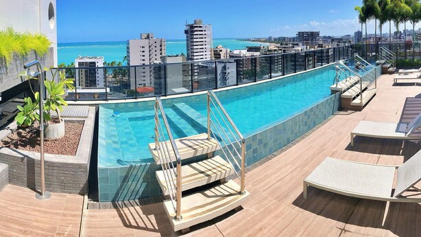 A rooftop pool with a city view, a glass barrier, and lounge chairs. - Vacanze - Time (Maceió)
