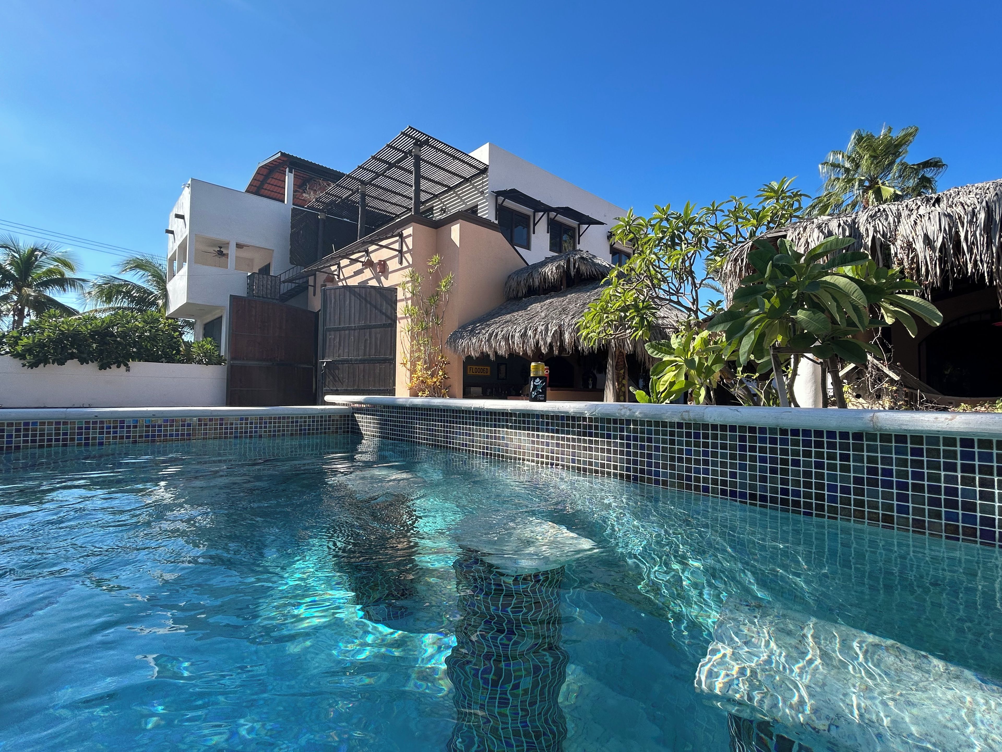 A swimming pool with clear blue water, surrounded by a white building with a thatched roof structure, palm trees, and a sign indicating a flooded area.
