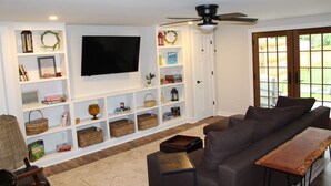 Uma sala de estar com sofá, televisão, estantes com livros e objetos decorativos, e vista para o exterior.