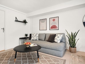 Una sala de estar con un sofá gris, una mesa de centro negra con un cuenco de fruta, una planta en maceta y cuadros en la pared.