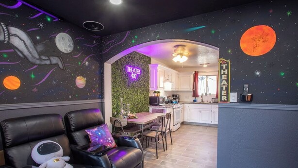 Une pièce avec un mur décoré sur le thème de l’espace, un canapé en cuir, une petite table et un coin cuisine avec des placards blancs et un néon.