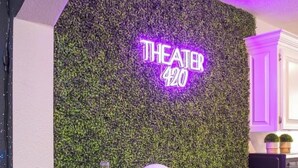 Un espace repas avec un mur vert orné d’un panneau néon indiquant « THÉÂTRE 420 », une table en bois et des chaises.