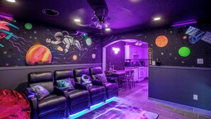 Une pièce comprenant un canapé, un fauteuil en forme de sac de haricots rouge, un ventilateur de plafond, un coin cuisine et un mur décoré sur le thème de l’espace.