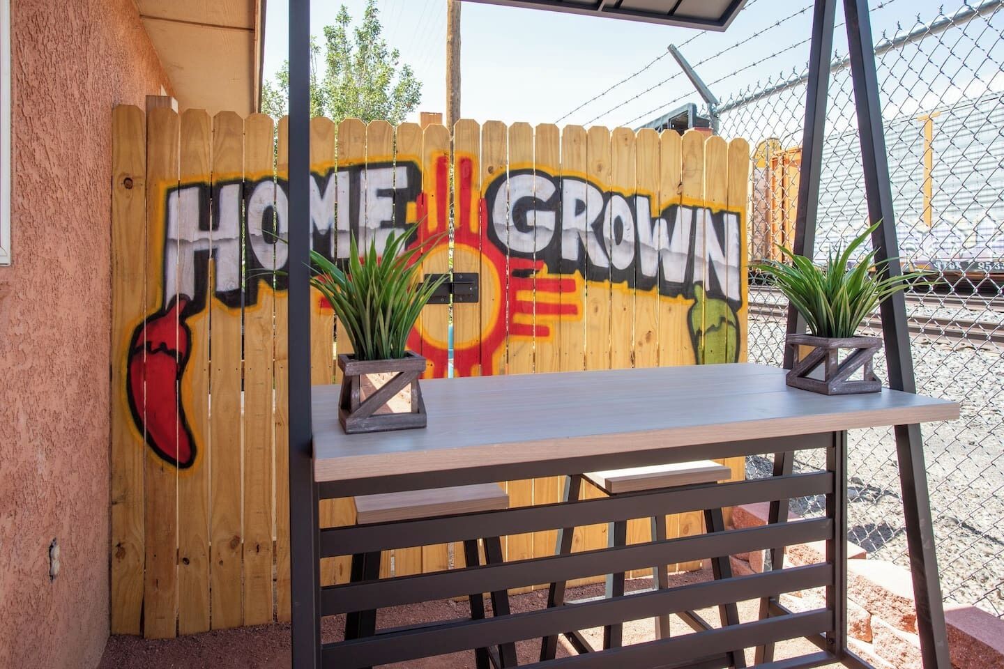 Una cerca de madera con grafiti que dice 'HOME GROWN', dos macetas y una mesa de metal.