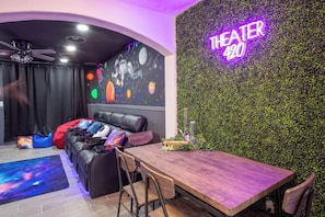 Ein Zimmer mit einem Holztisch, schwarzen Ledersitzmöbeln, einem leuchtenden Schild mit der Aufschrift 'THEATER 420' und einer Wand, die mit Grünpflanzen bedeckt ist.