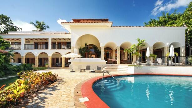 A pool area with a red border, lounge chairs, and a building with multiple balconies and arches. - Stunning Ocean View Villa (La Romana)