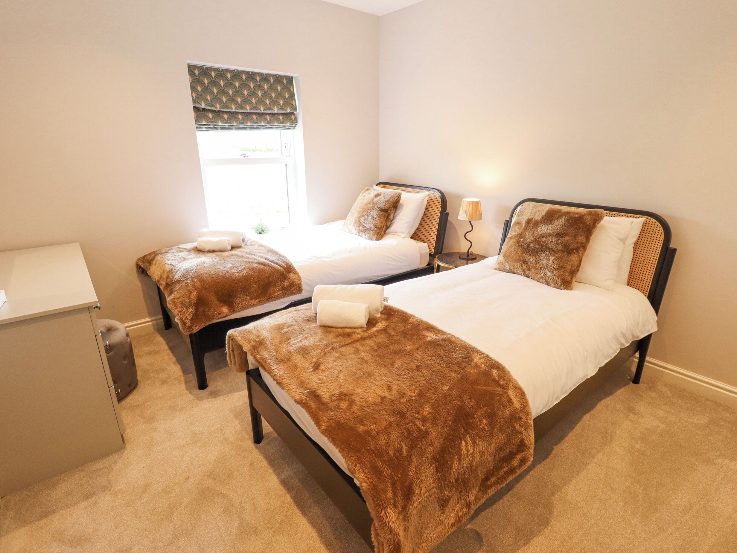 A hotel room with two single beds, a small desk, a window with a patterned blind, and a lamp on the nightstand.