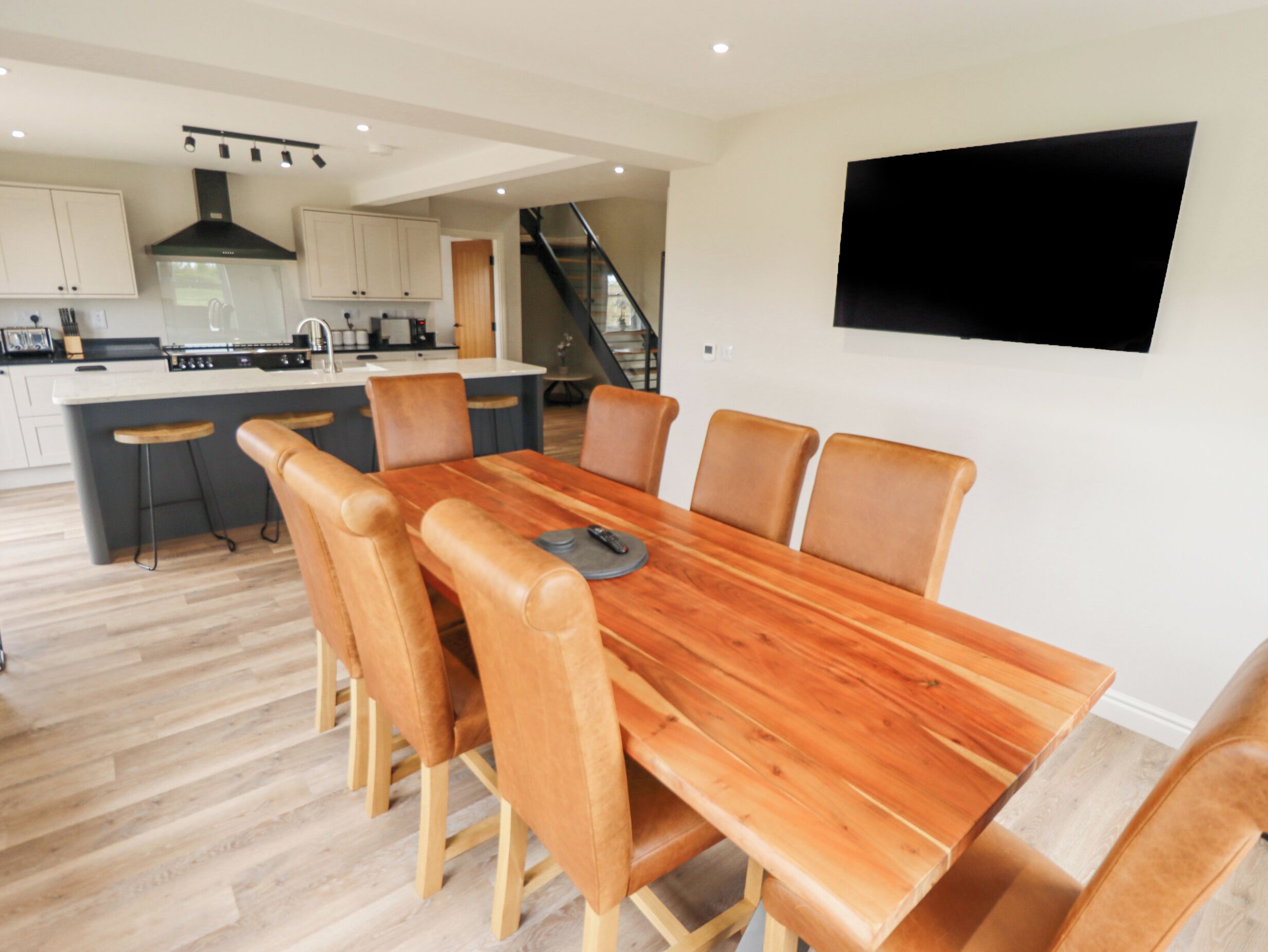 Une cuisine moderne dotée d’un îlot central, un coin repas avec une table en bois et des chaises en cuir, et un escalier menant à un niveau supérieur.