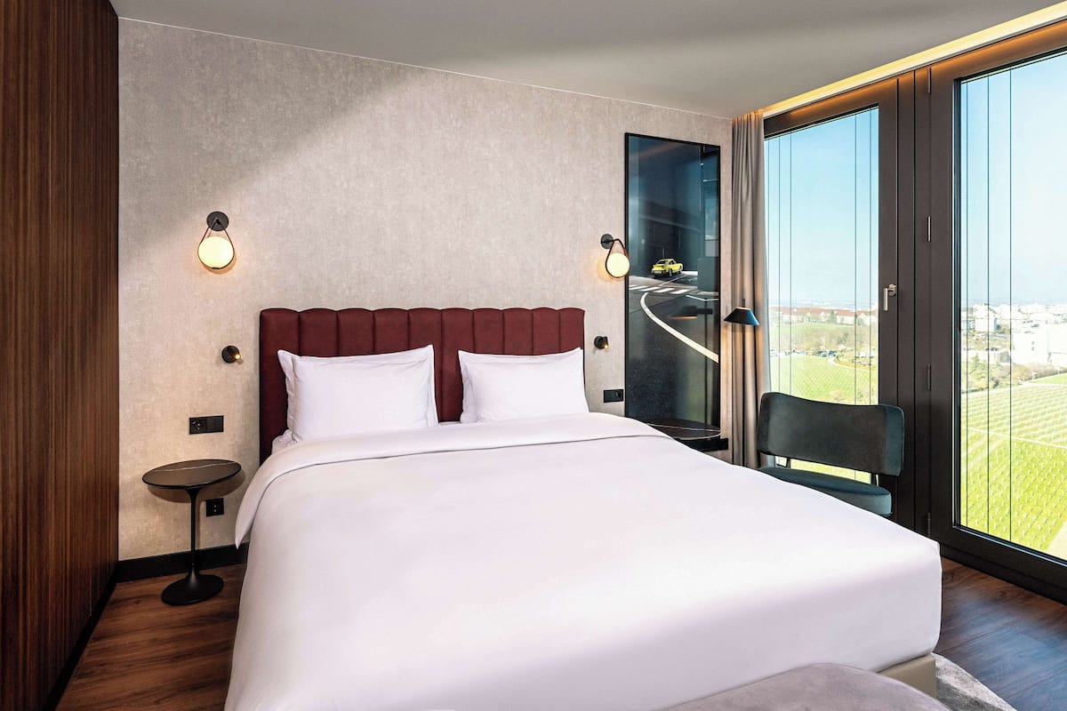 A modern hotel room with a large bed, two wall-mounted lights, a small side table, and a view of a cityscape through large windows.